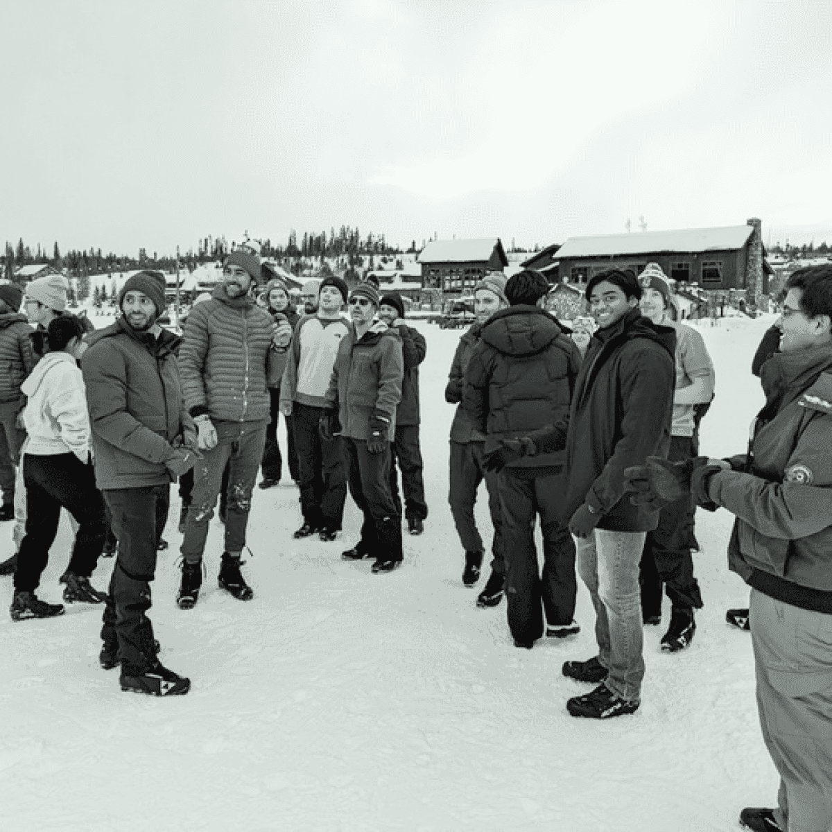 A group of people standing together outdoors in a snowy landscape, dressed in winter clothing, with buildings and trees in the background.