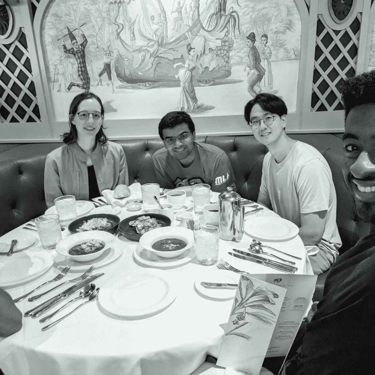 A group of people seated around a round table in a restaurant, smiling at the camera, with plates and cutlery set for a meal.