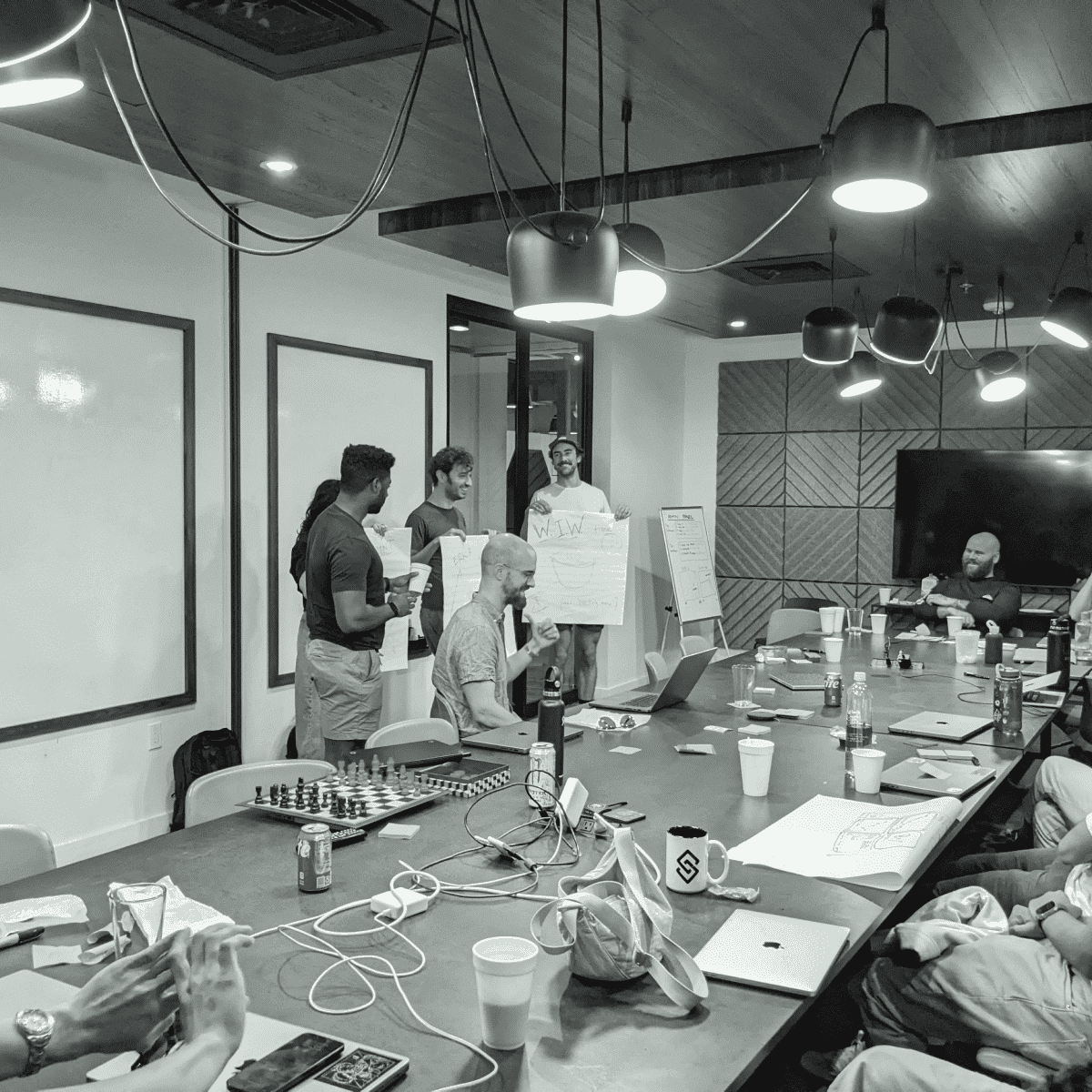 A group of people in a conference room, some standing and presenting with flip charts, while others are seated around a table covered with laptops, papers, and coffee cups.