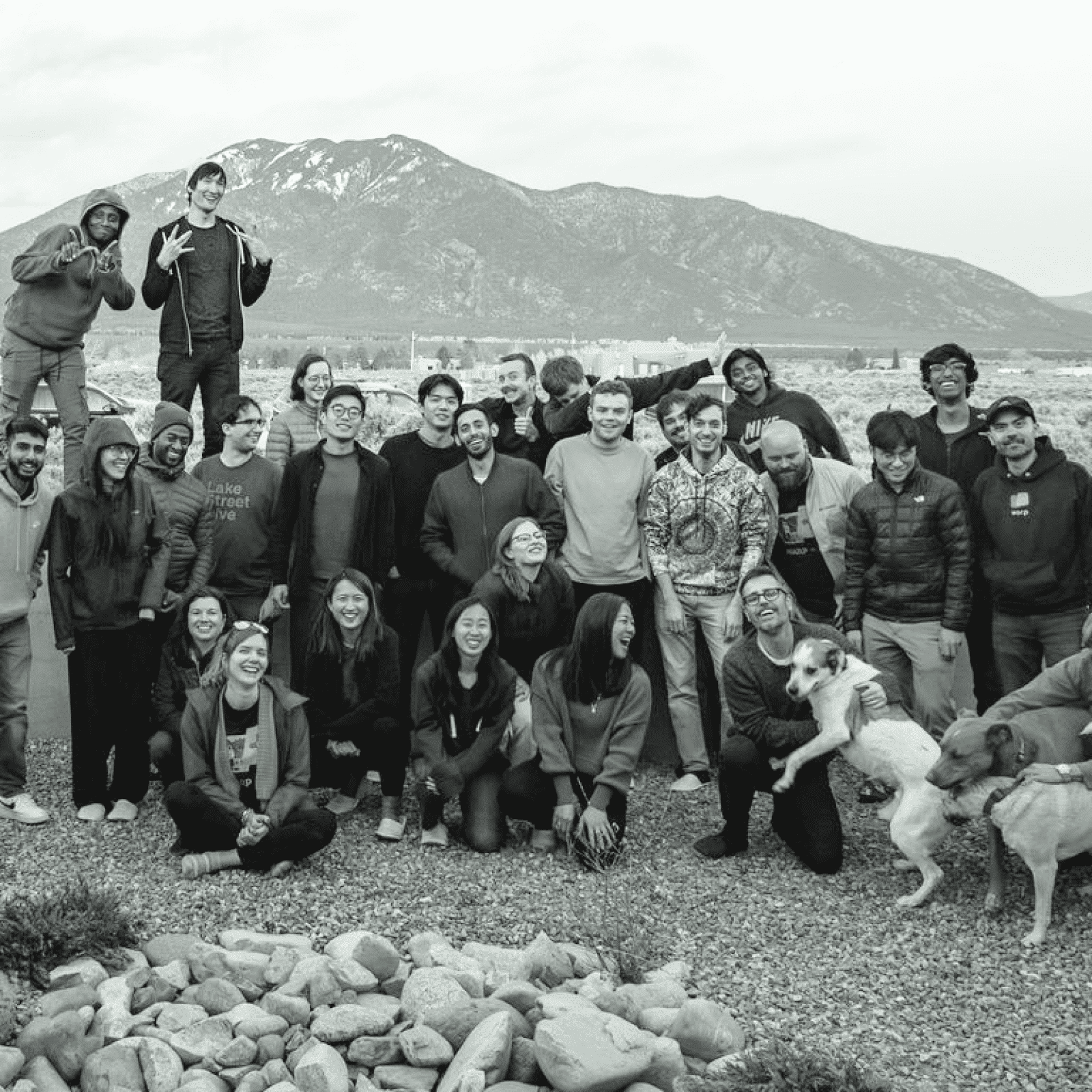 A large group of people posing together outdoors with mountains in the background, some individuals standing, some kneeling, and a few holding dogs.