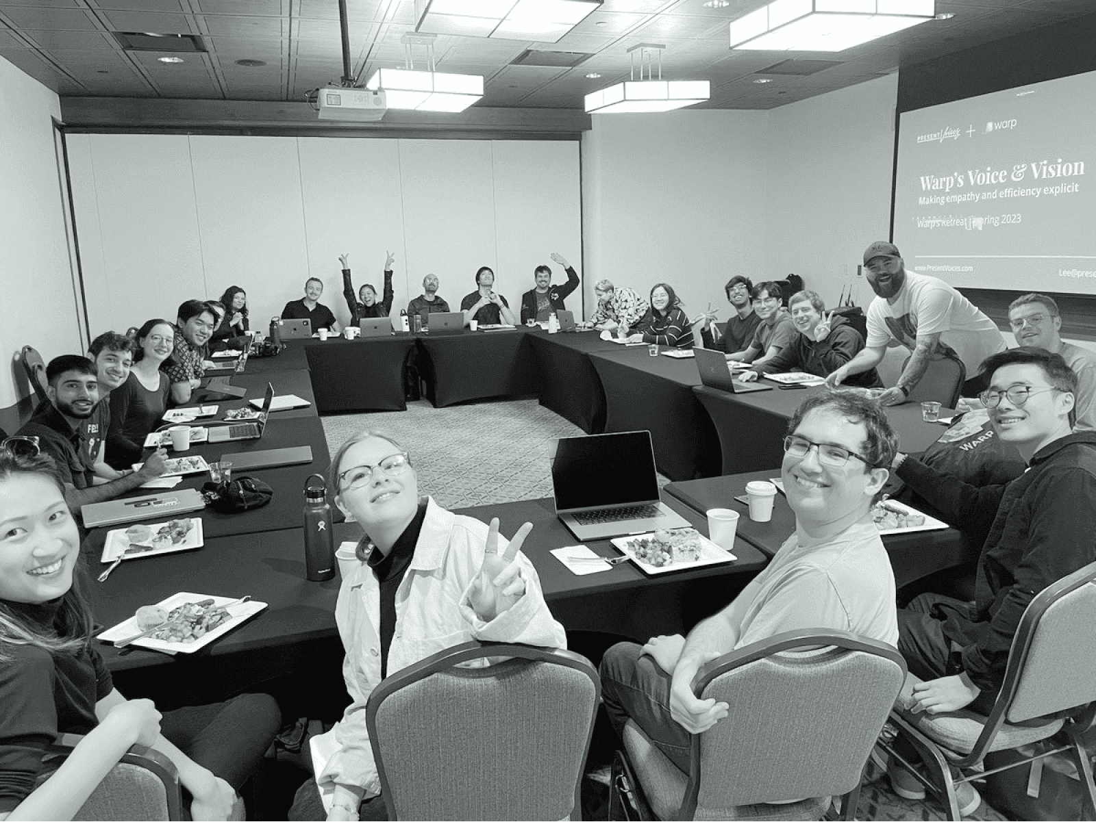 A large group of people sitting around a U-shaped conference table in a meeting room, smiling and making gestures towards the camera.